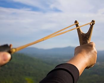 A person aiming a slingshot outdoors with a scenic landscape in the background.